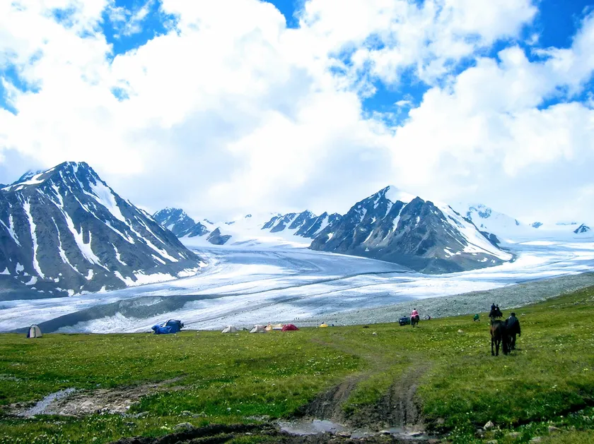 A camp and riders on a green meadow below a glacier between snow peaks
