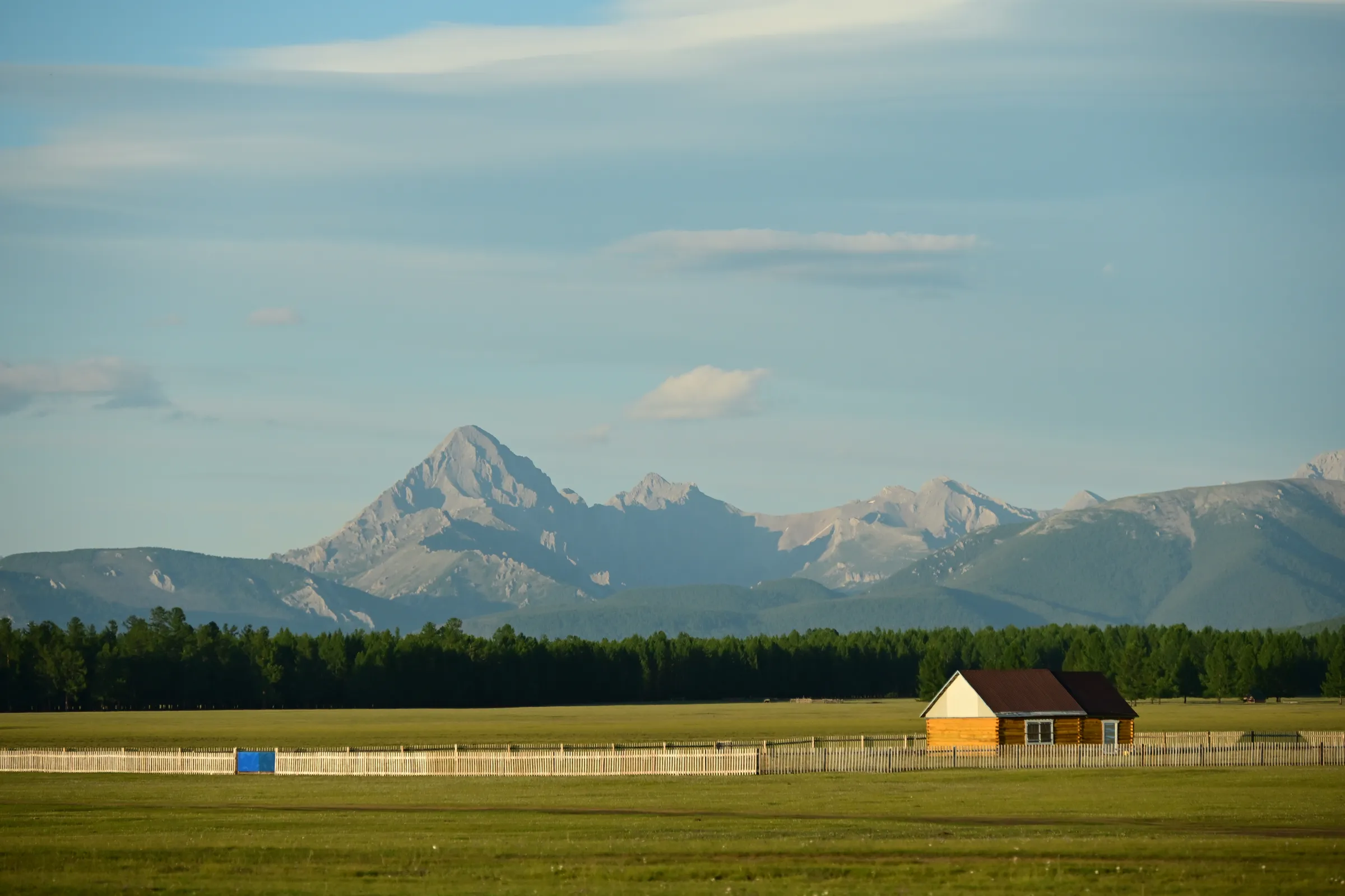 A log cabin behind a white fence on open grassland, dark forest and granite peaks beyond
