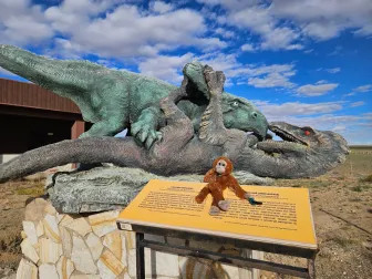 A plush monkey sitting on the sign of a fighting-dinosaurs statue in the Gobi