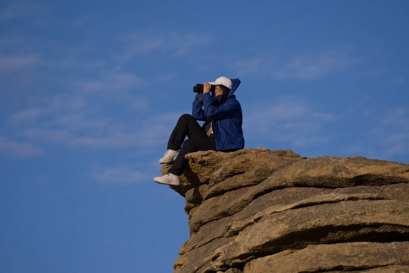 A traveller with binoculars sitting on the edge of a granite ledge against a blue sky