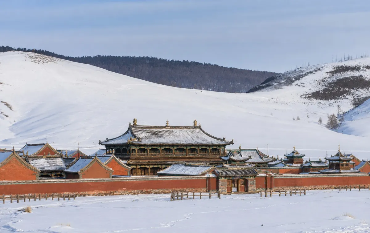 A monastery of tiered roofs behind a red wall, under snow, with white hills behind