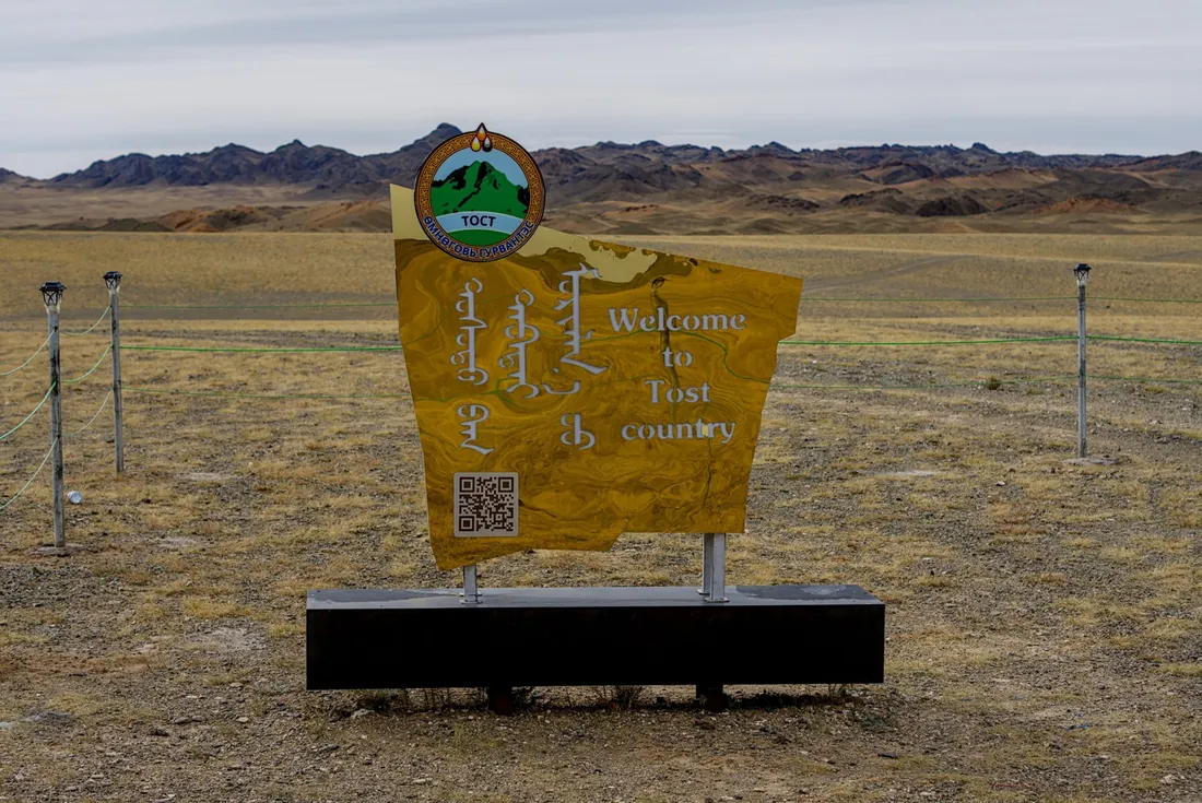 The Welcome to Tost country sign on the gravel plain, with the Tost Mountains behind