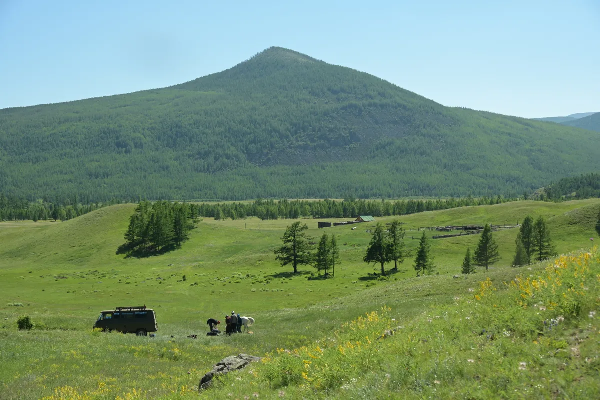 A van at the end of a track, riders loading a pack horse below a forested mountain
